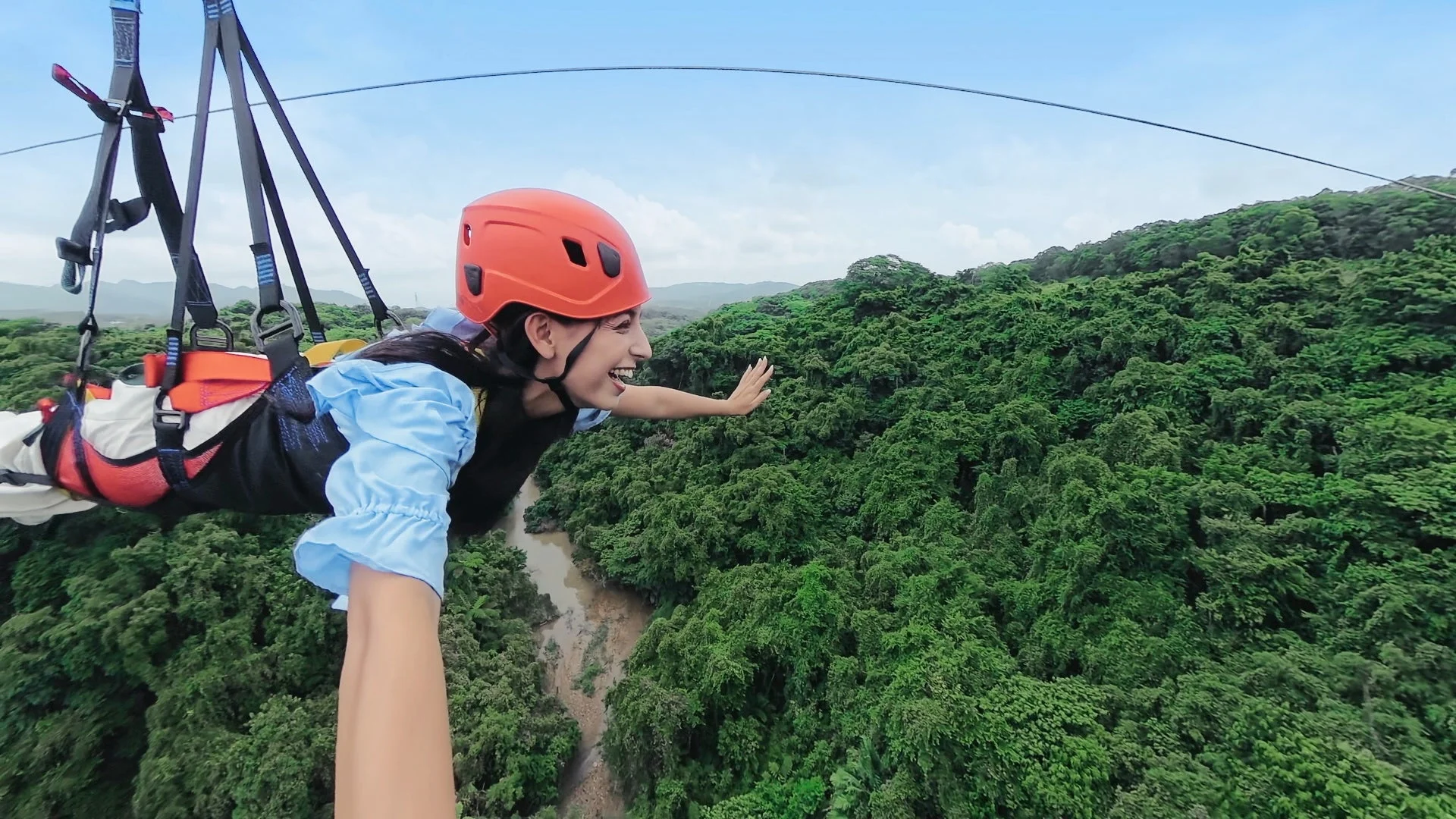 緑豊かな森林と川の上を、オレンジ色のヘルメットとハーネスを装着した女性がジップラインで滑空しています