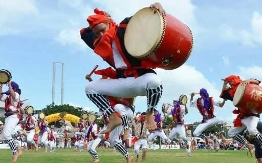 青空の下、広々とした芝生の上で、多くの人々が伝統的な衣装を身につけ、躍動感あふれるエイサーを披露しています。