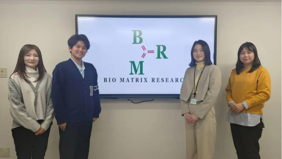 会議室のような場所で、スクリーンに「BIO MATRIX RESEARCH」のロゴが表示されており、男女4人が笑顔で立っている様子です。