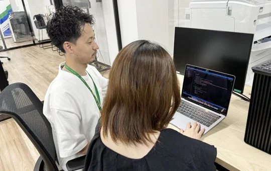 オフィスで男性と女性が並んで座っており、女性がラップトップパソコンでコードのようなものを表示させながら作業しています