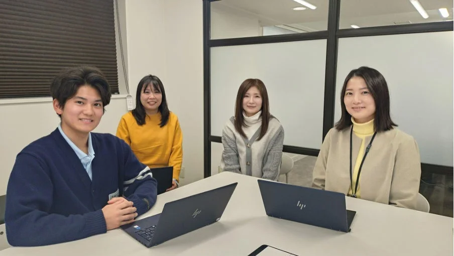 オフィスまたは会議室で、男女4人がテーブルを囲んで座っています。皆笑顔で、テーブルにはノートパソコンが置かれており、和やかな雰囲気で会議や共同作業をしている様子です。