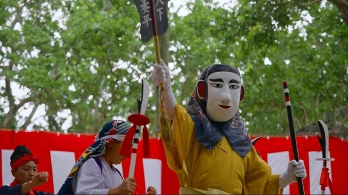 伝統的な黄色い衣装と大きな仮面をつけた人物が屋外でパフォーマンスを行っています。手に持った旗には「天下太平」と書かれており、日本の文化的な祭りや芸能の様子を捉えた一枚です。