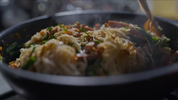 黒いフライパンで麺料理が調理されている様子。湯気が立ち上り、麺、ソーセージ、野菜、きのこなどが炒められている、温かい食事の準備風景です。