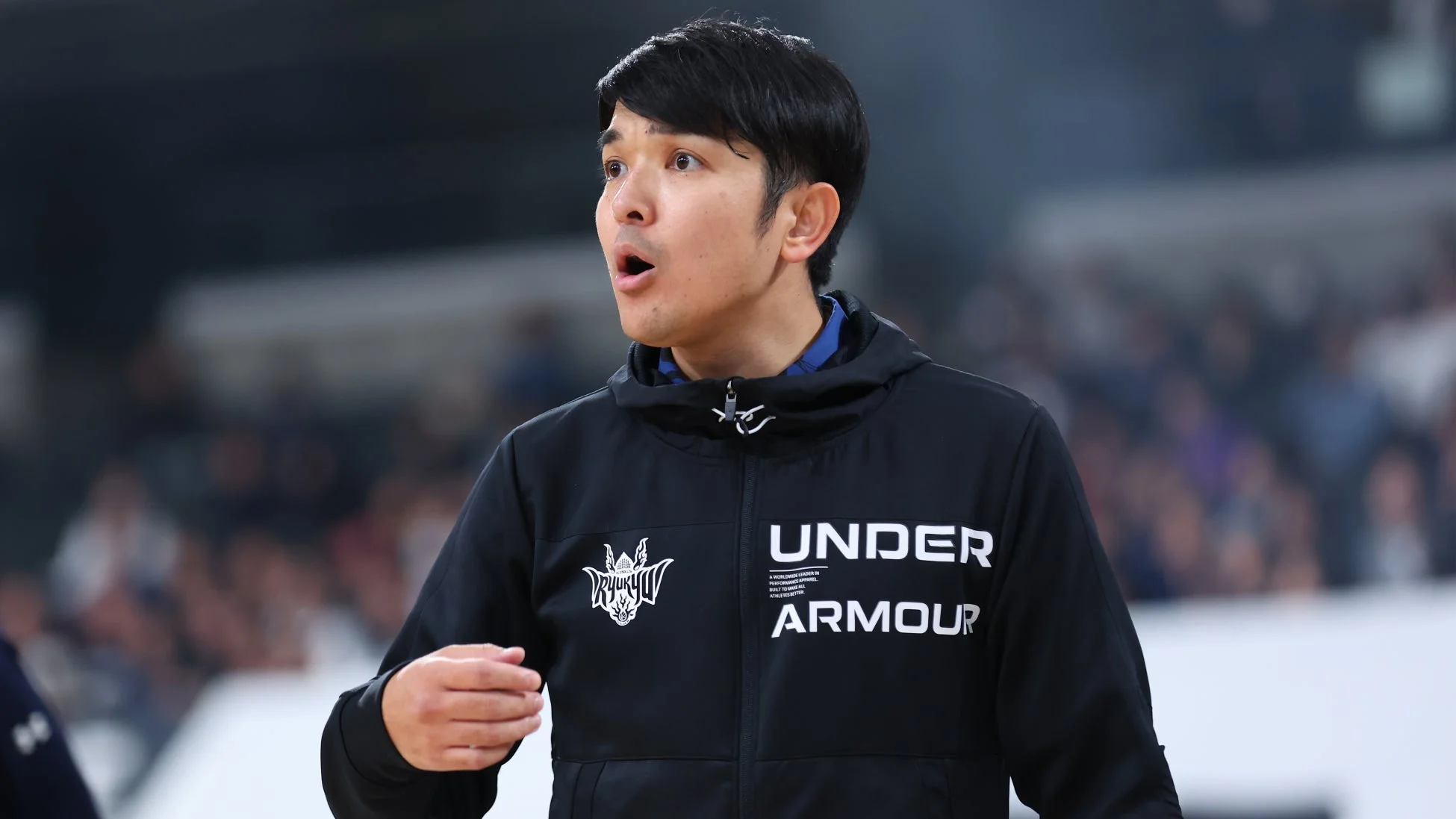 黒いアンダーアーマーのジャケットを着た男性が、琉球のロゴをつけ、スポーツイベント中に何かを指示しているかのような真剣な表情