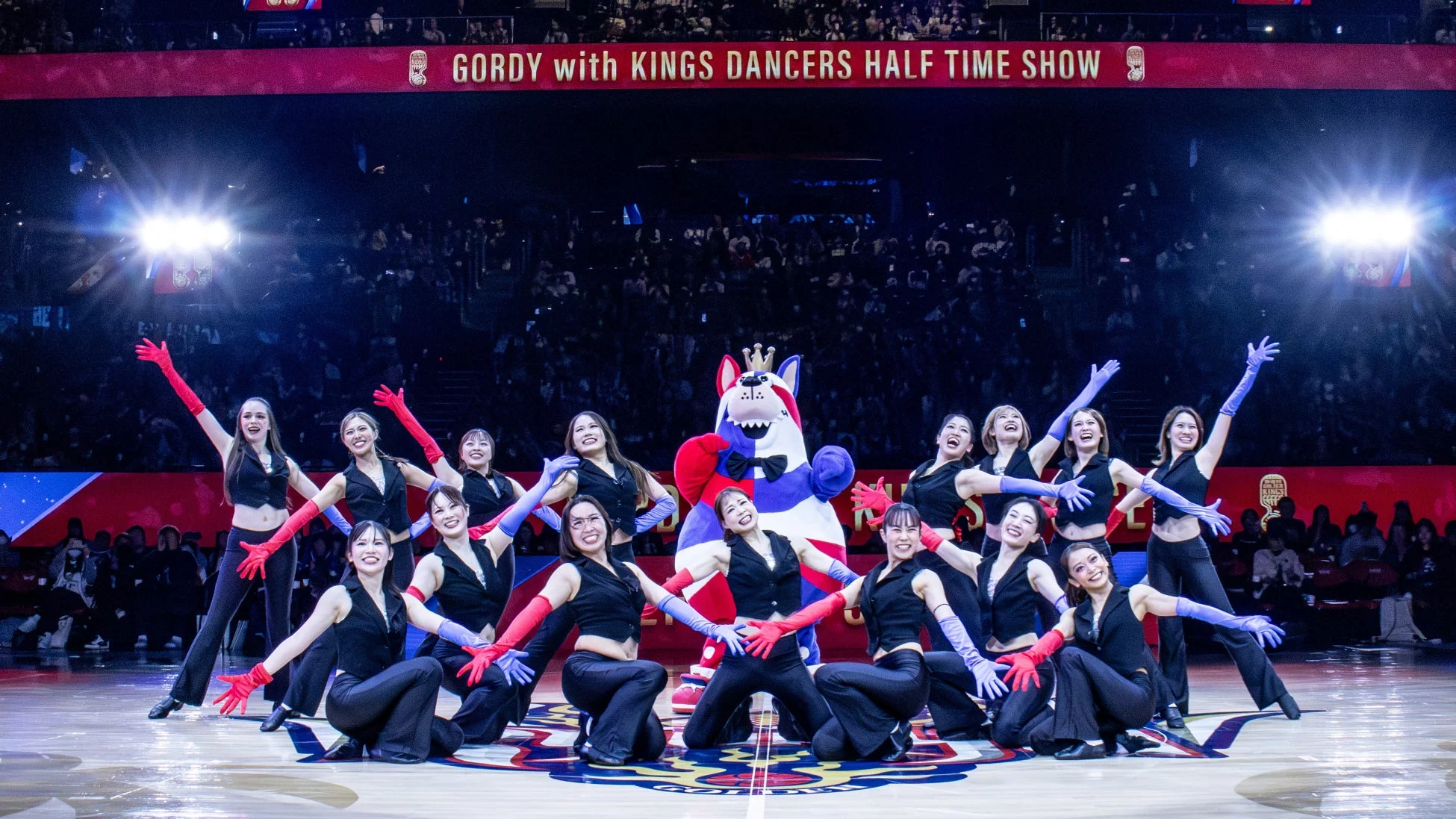 アリーナで行われているハーフタイムショーで、カラフルな手袋を着用したダンサーたちがマスコットキャラクターと一緒にポーズをとっています