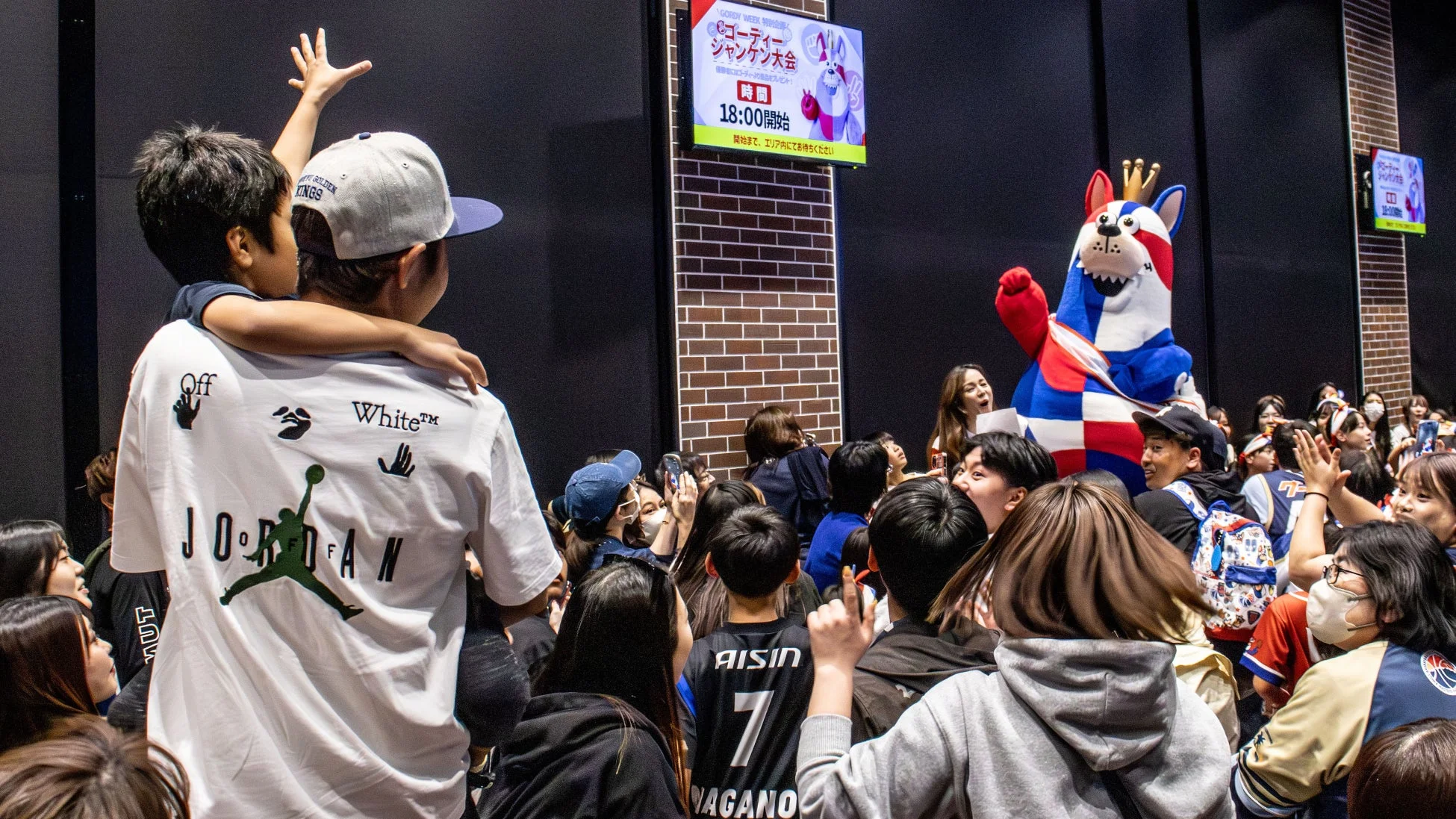 多くの観客が屋内に集まり、ステージ上のマスコットキャラクター「ゴーディー」とスクリーンに注目しています