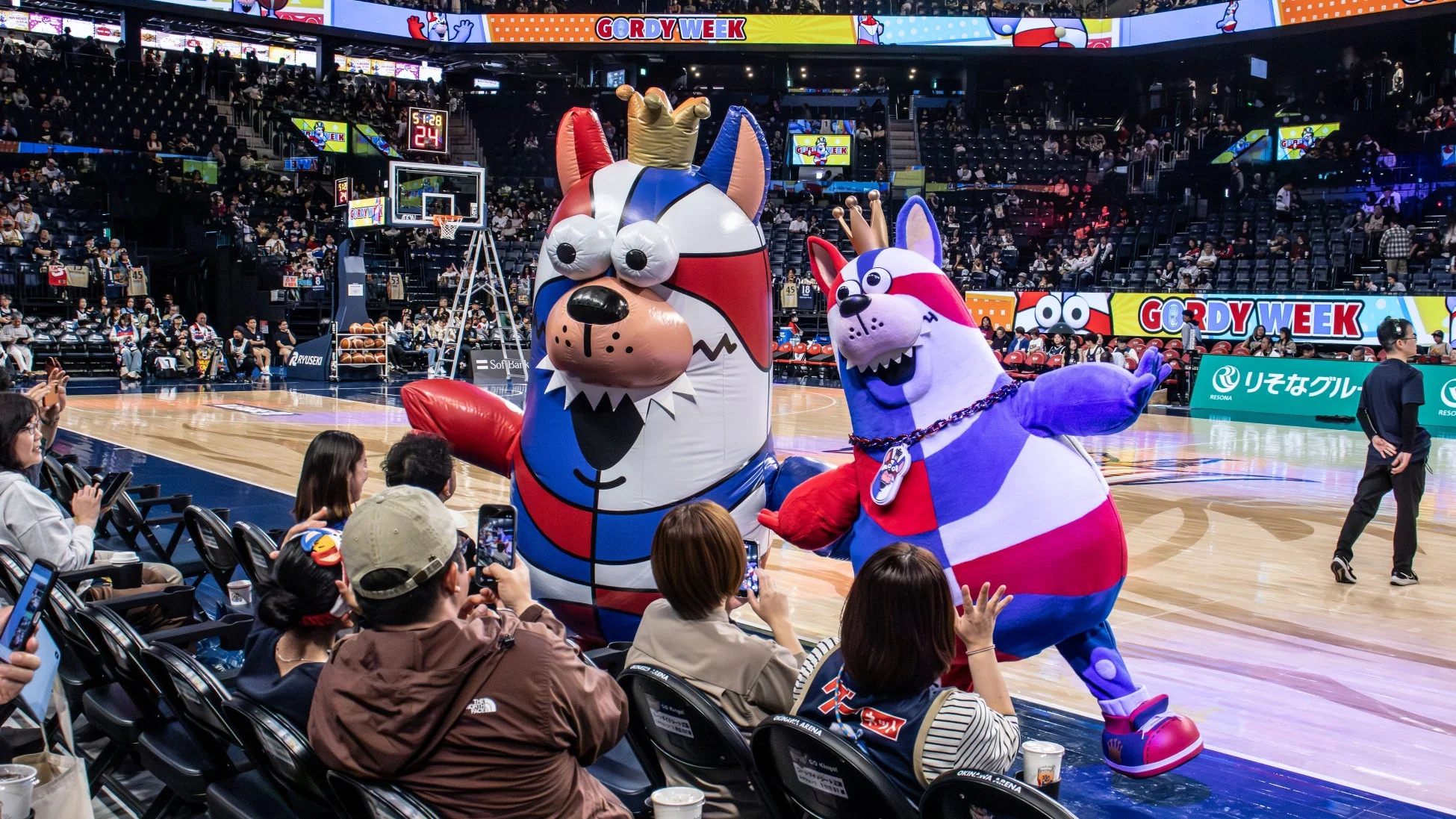 バスケットボールのアリーナで、巨大なマスコットキャラクター2体が観客の前でパフォーマンスをしている様子