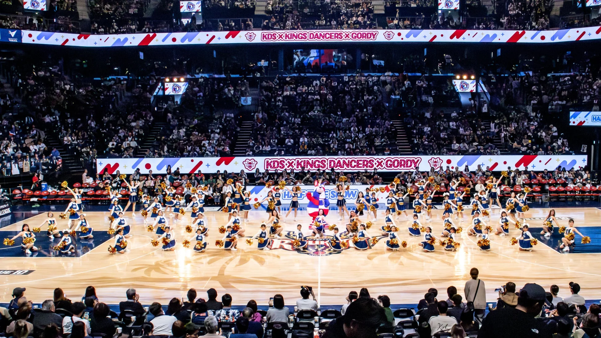 バスケットボールコートで、多数のチアリーダーがマスコットと共に華やかなダンスパフォーマンスを披露している
