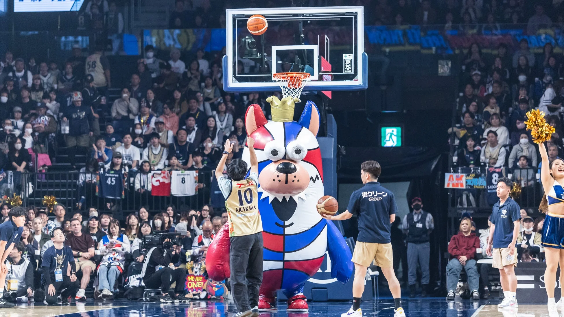 バスケットボールの試合会場で、選手が巨大なマスコットキャラクターの傍らでシュートを放っている瞬間