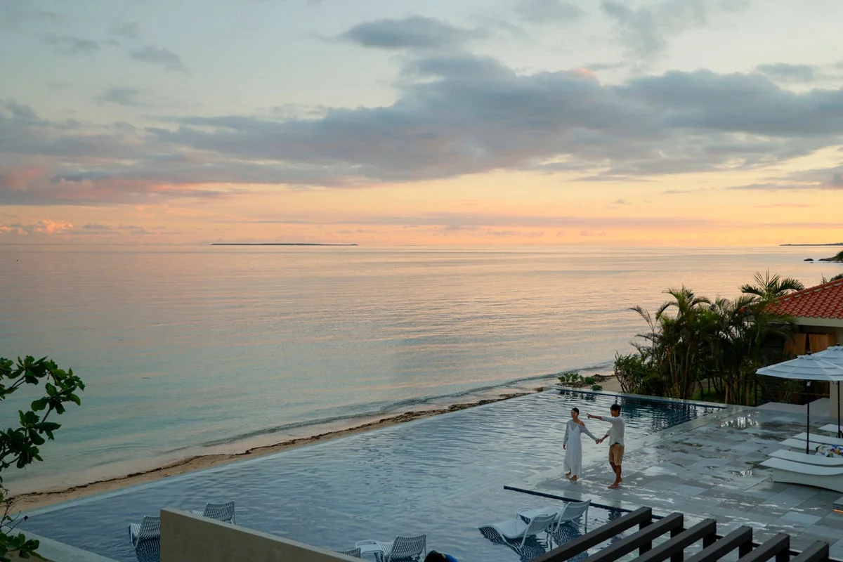夕焼けに染まる海とインフィニティプールを背景に、手をつないで立つカップル