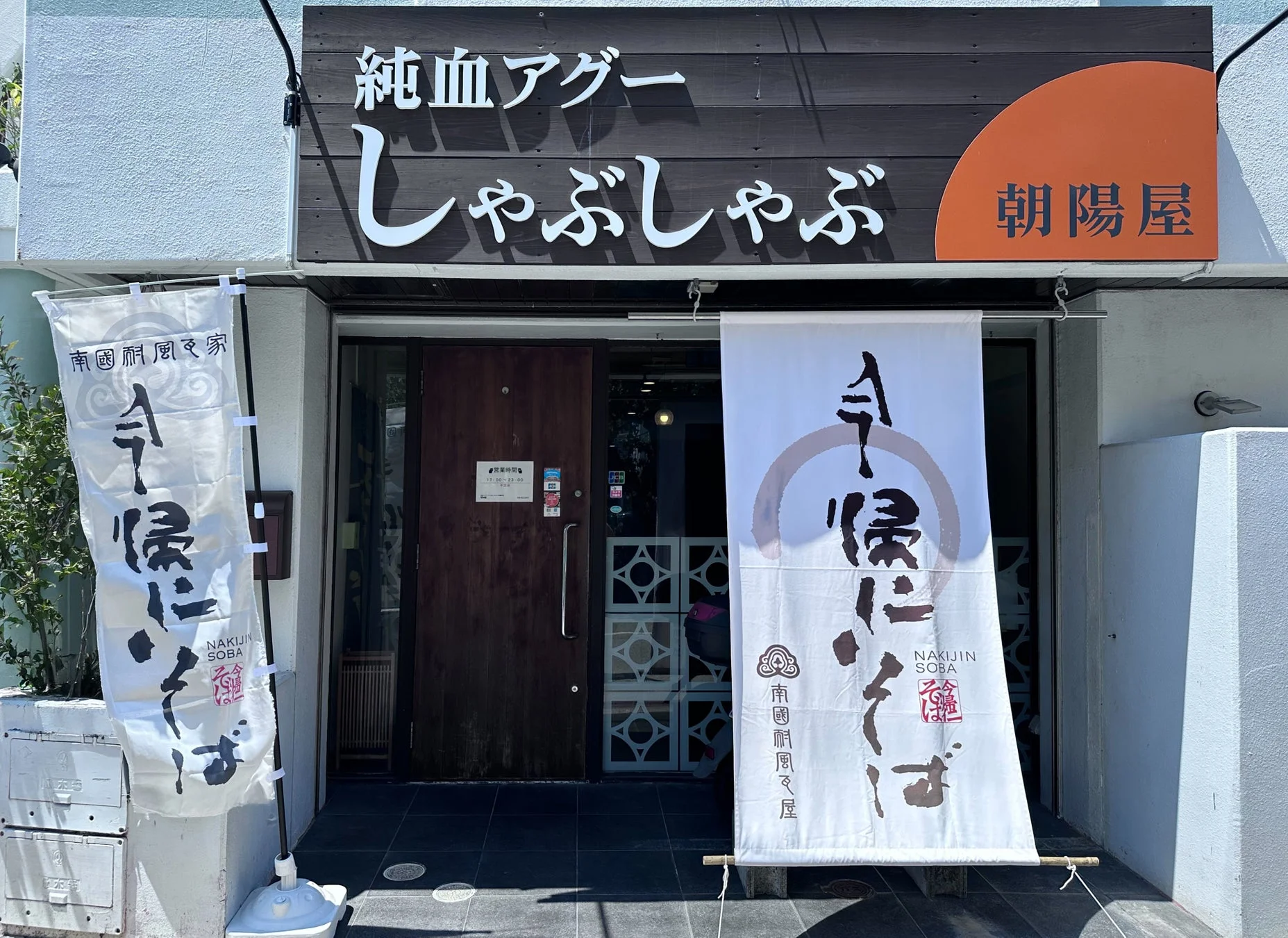 「純血アグーしゃぶしゃぶ 朝陽屋」と「今帰仁そば」を提供する飲食店の外観。店頭には今帰仁そばののぼり旗が2本立てられ、営業時間は13:00から23:00と表示されている。沖縄料理を提供する店舗の様子。