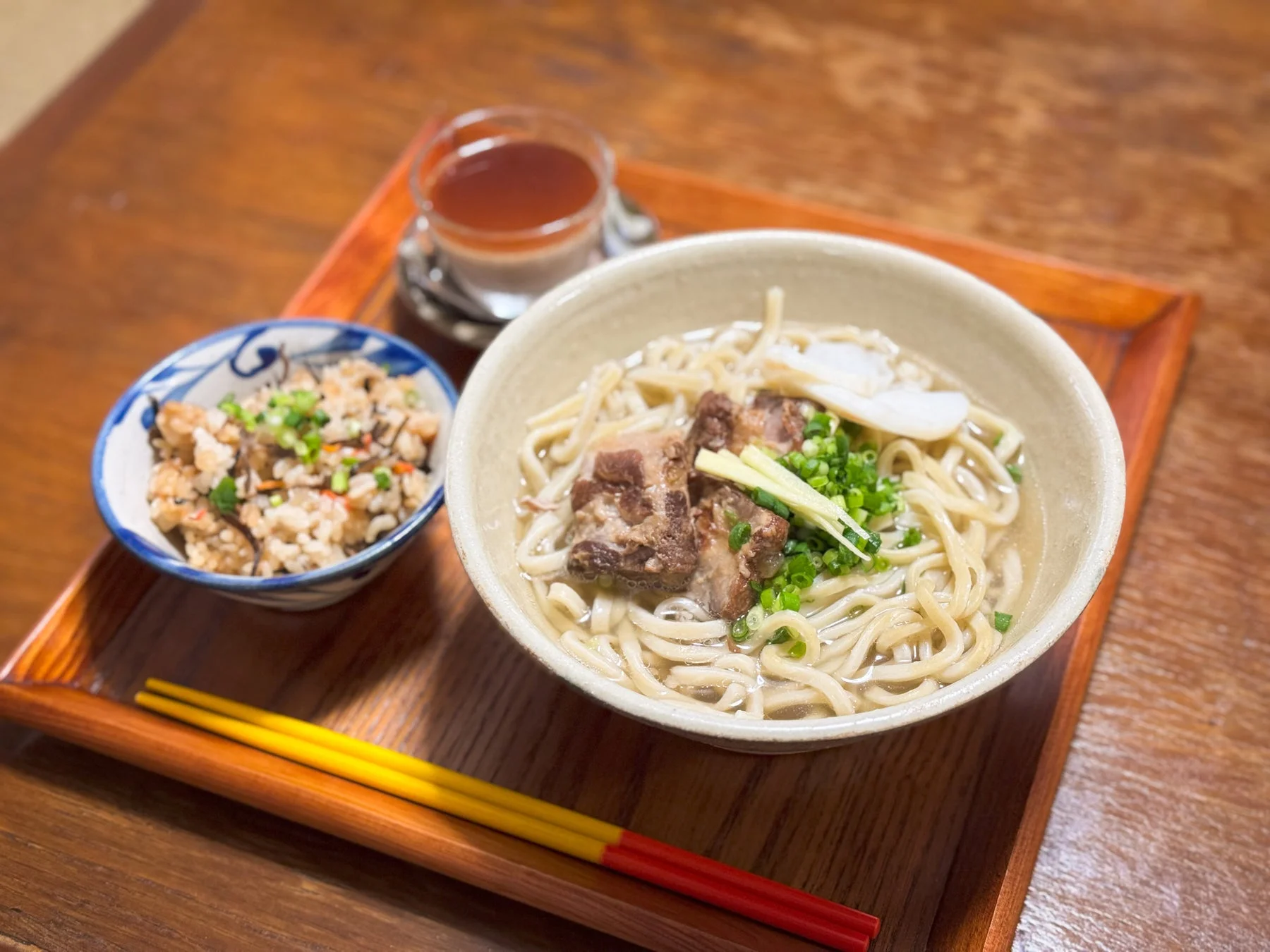 木のトレーに盛られた沖縄そばとじゅーしーの定食です。豚肉の角煮やネギがのった温かいそばと、彩り豊かな炊き込みご飯が食欲をそそります。飲み物も添えられ、バランスの取れた和食のセットです。