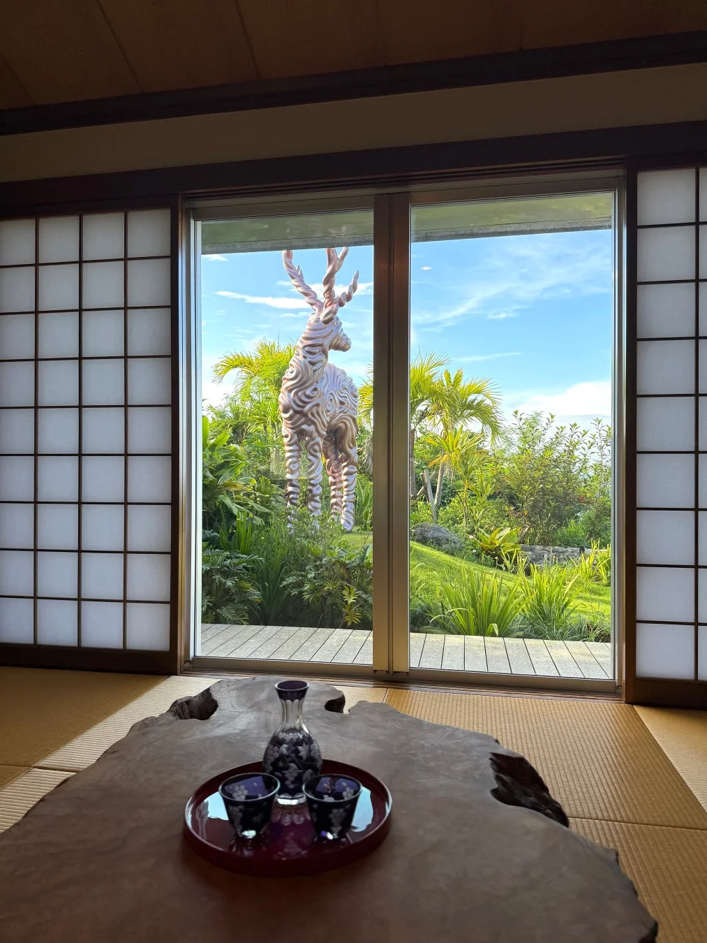 畳と障子のある和室から、広々とした庭園を望む風景。庭にはヤシの木が生い茂り、流線型の模様が特徴的な鹿の現代アート彫刻が際立っている。手前の木製テーブルには日本酒のセットが置かれ、晴れた日の穏やかな雰囲気が漂う。