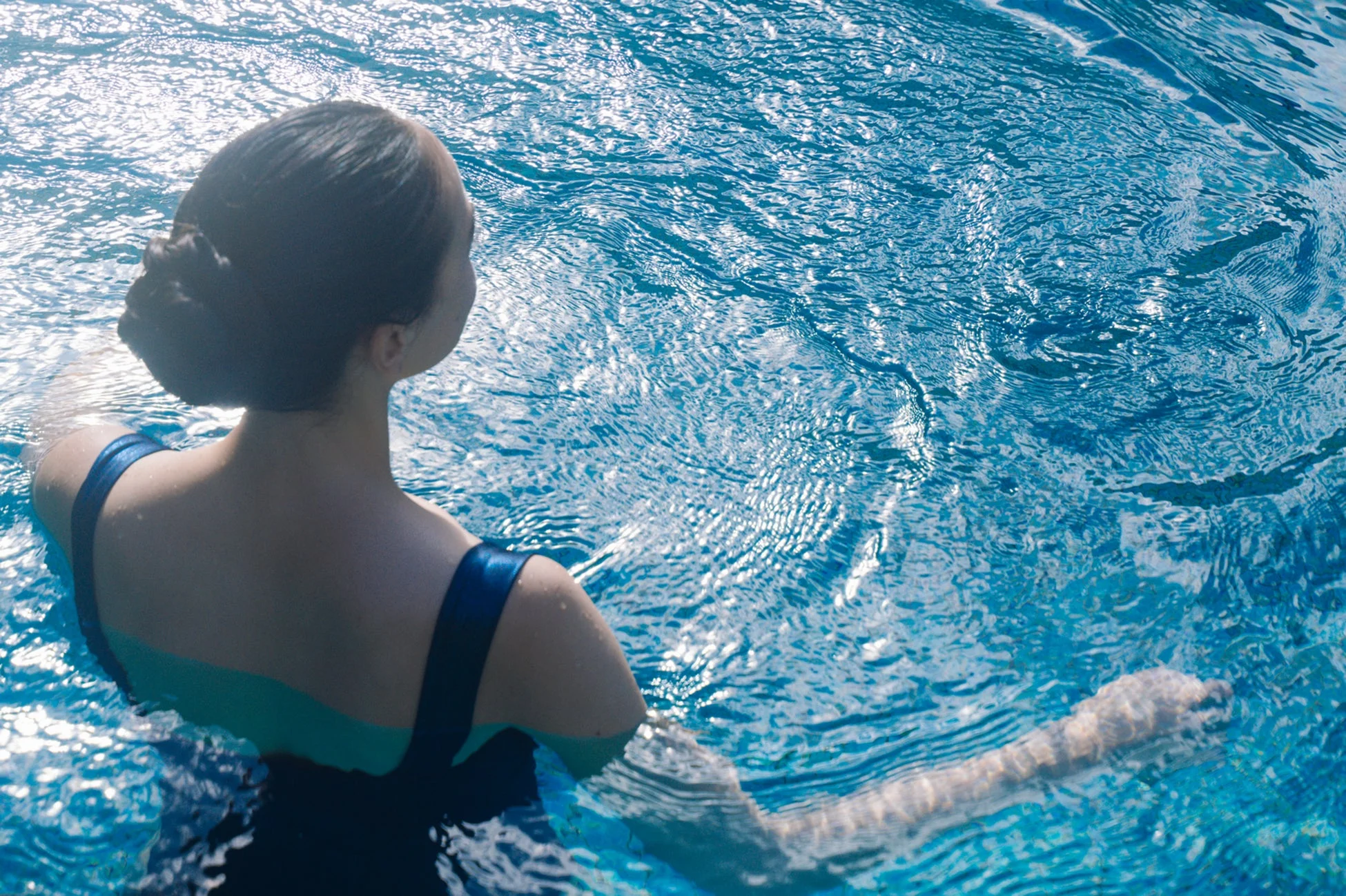 青い水面が輝くプールで水に浸かる女性の後ろ姿