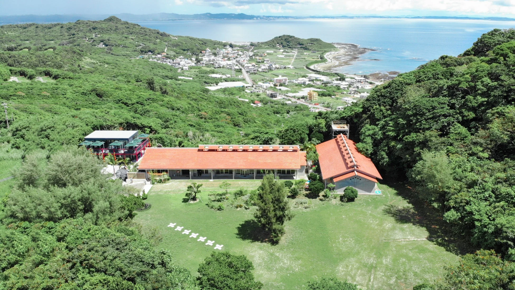 上空から撮影された、緑豊かな丘陵地帯に建つ複数の建物と、その向こうに広がる青い海と海岸沿いの町並みが見える美しい風景。晴れた日の開放的な景色が特徴です。