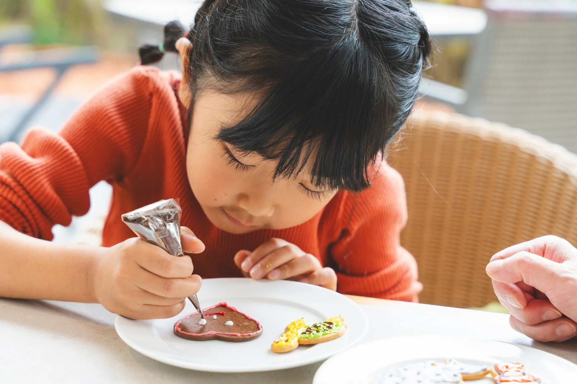 オレンジ色のセーターを着た小さな女の子が、真剣な表情でクッキーにデコレーションをしている様子。絞り袋を使い、ハート型のクッキーに白いアイシングで模様を描いています。親子でのお菓子作りを楽しんでいる瞬間を捉えた写真です。