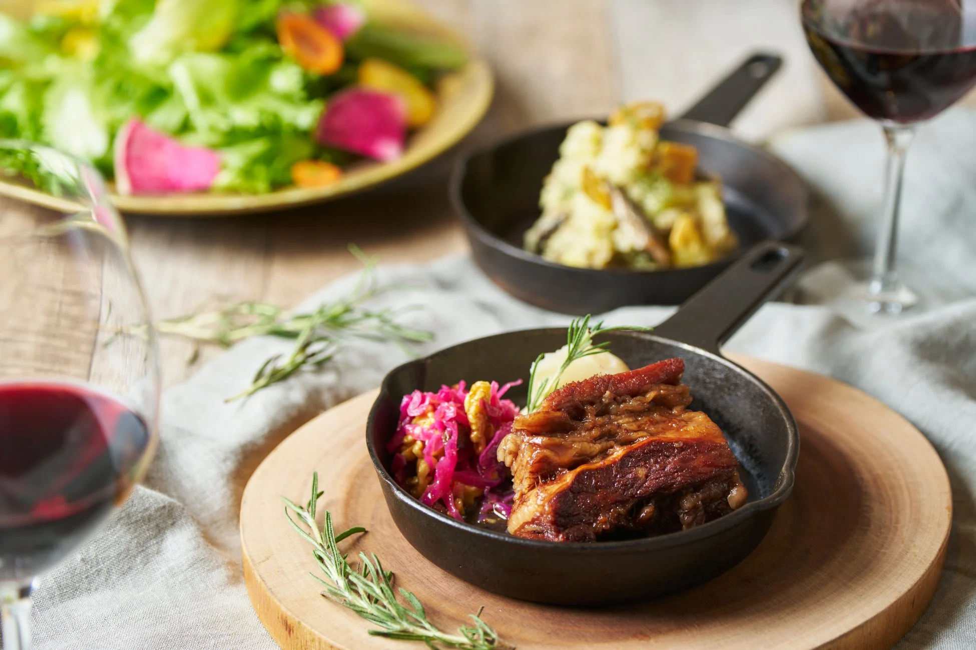 スキレットに盛り付けられた煮込み肉料理と赤ワイン、サイドディッシュのポテトサラダや彩り豊かなサラダが並ぶおしゃれなディナー風景。温かい食事が楽しげな雰囲気を醸し出しています。