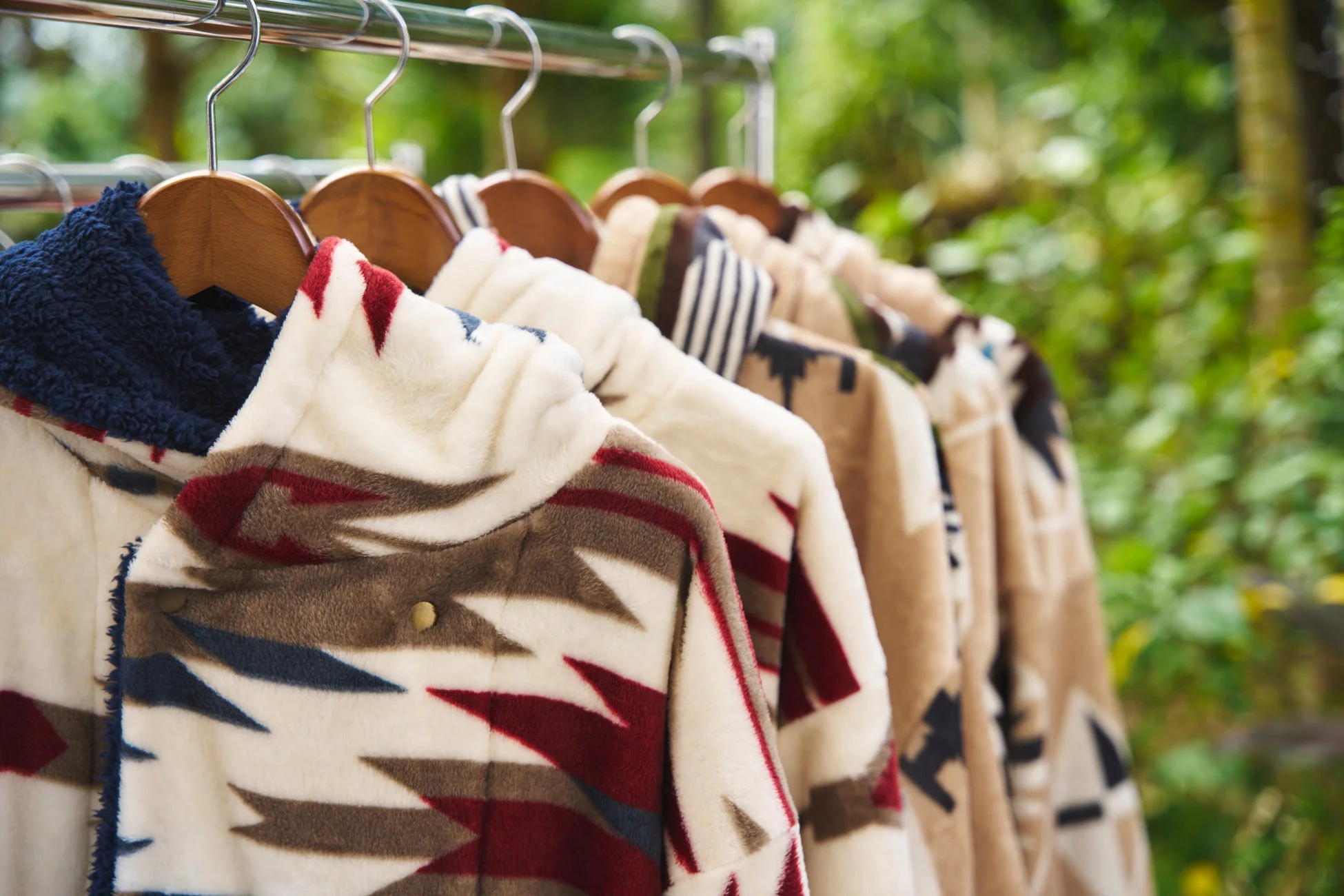 ネイティブ柄のような幾何学模様が特徴のフリースジャケットが、屋外のハンガーラックに複数掛けられている様子。暖かそうなアウターが並び、背景には緑豊かな自然がぼやけて見える。
