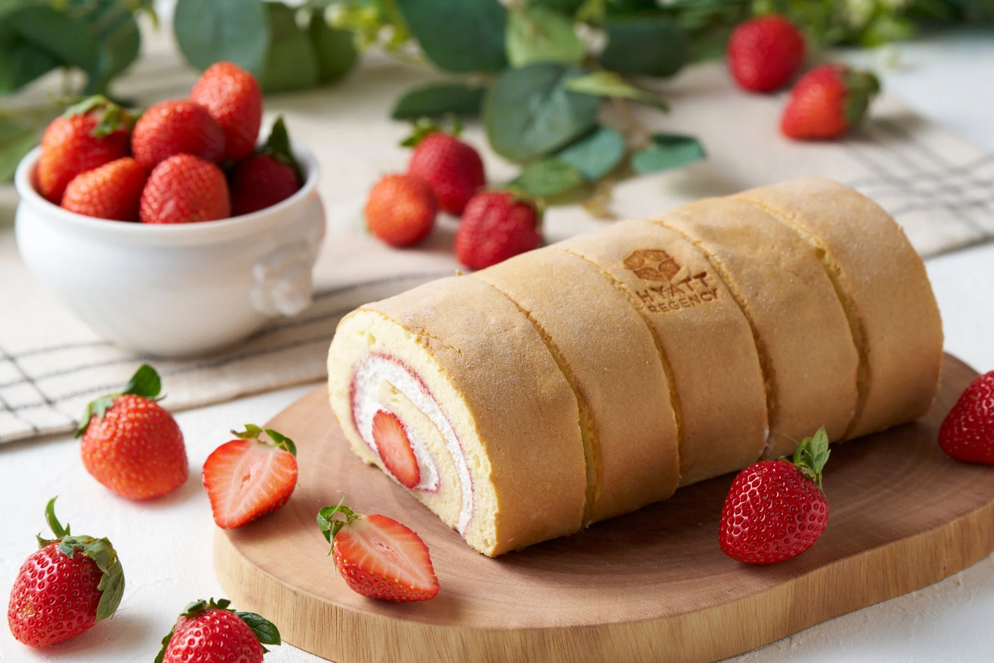 「HYATT REGENCY」のロゴが入ったロールケーキが木製のボードに置かれ、周りには新鮮なイチゴが散りばめられています。クリームとイチゴが美しい断面を見せる、美味しそうなデザートの画像です。
