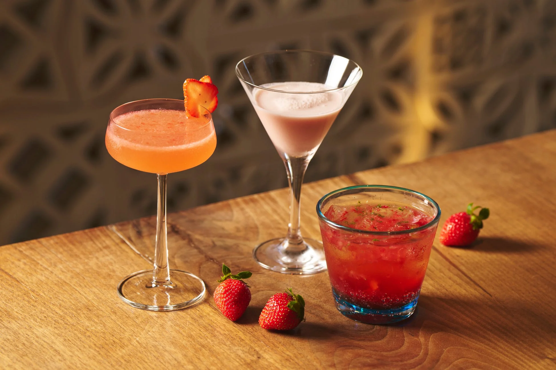木製のテーブルに、イチゴを添えた3種類のカクテルが並べられています。オレンジ色のカクテル、ピンク色のクリーミーなカクテル、そしてイチゴがたっぷり入った赤いソーダのようなドリンクがあり、新鮮なイチゴも添えられています。