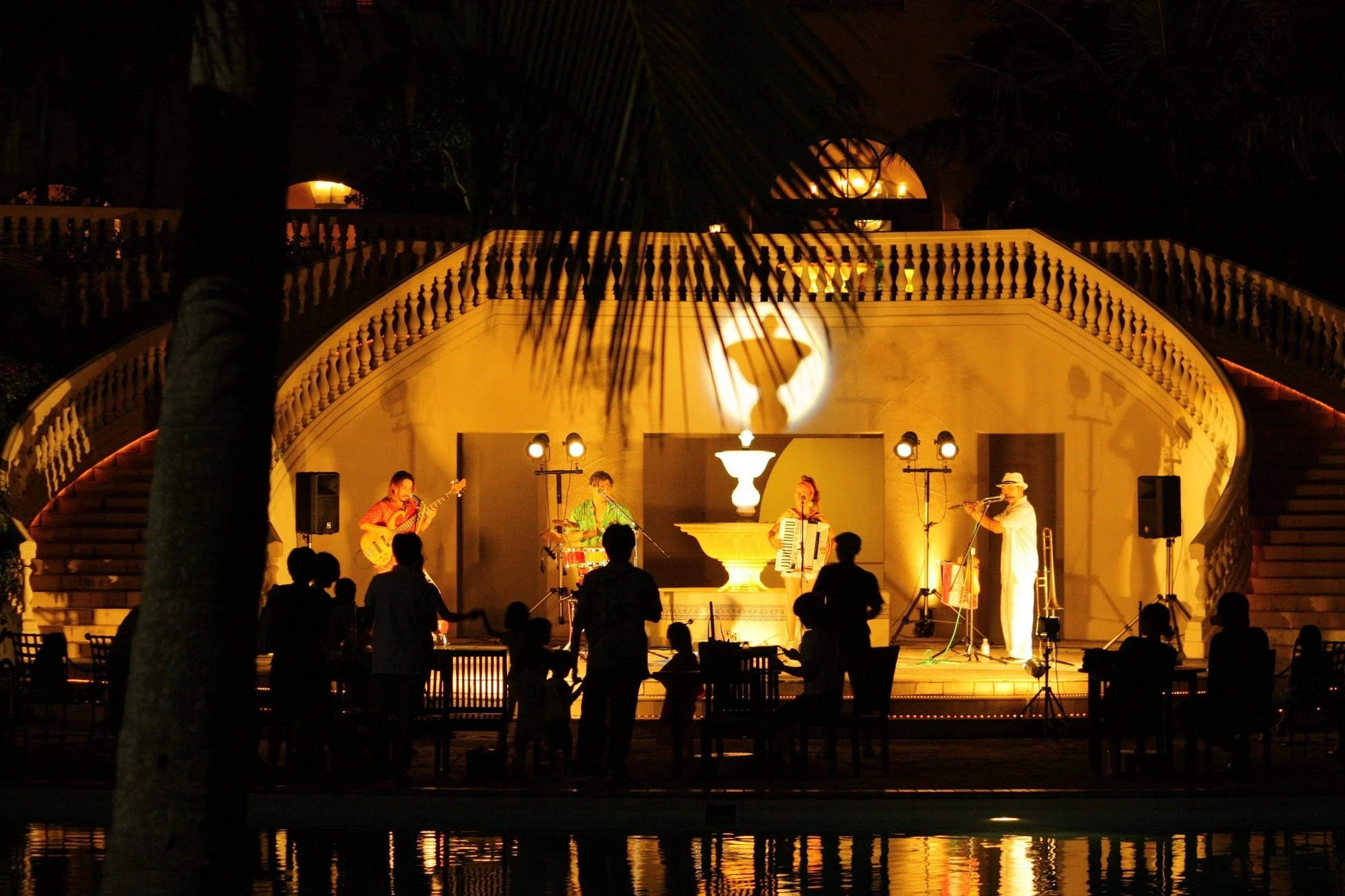 夜景、屋外ライブ、バンド演奏、音楽、コンサート、観客、ステージ、ライトアップ、ヤシの木、リゾート、プール、夜のイベント、エンターテイメント、楽器、ベース、アコーディオン、フルート、ドラム、階段、反射