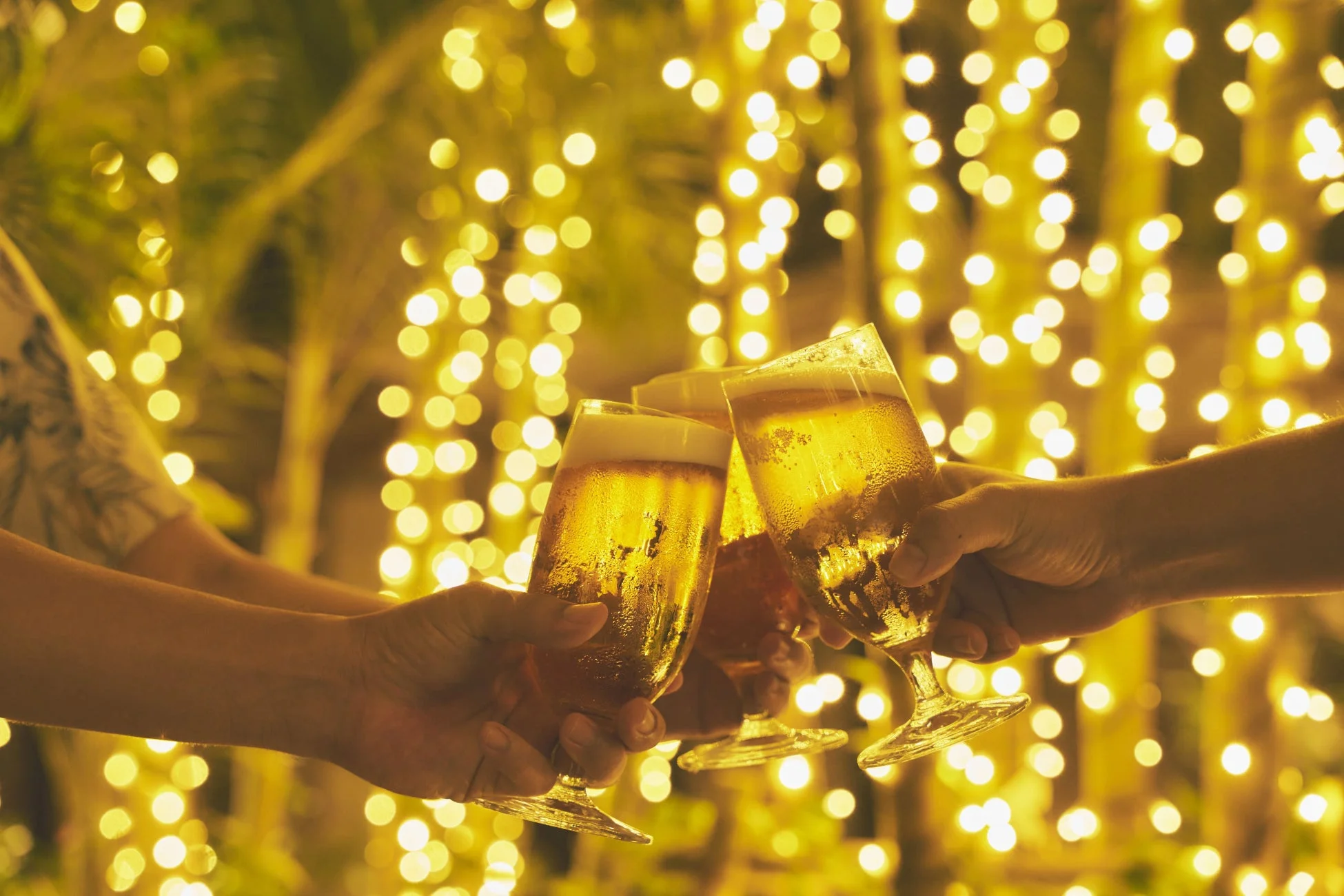 冷えたビールグラスを手に乾杯する複数の人々が写っています。背景には温かい光のイルミネーションが輝き、祝祭的で楽しい雰囲気を醸し出しています。友人や仲間との集まりの一コマのようです。