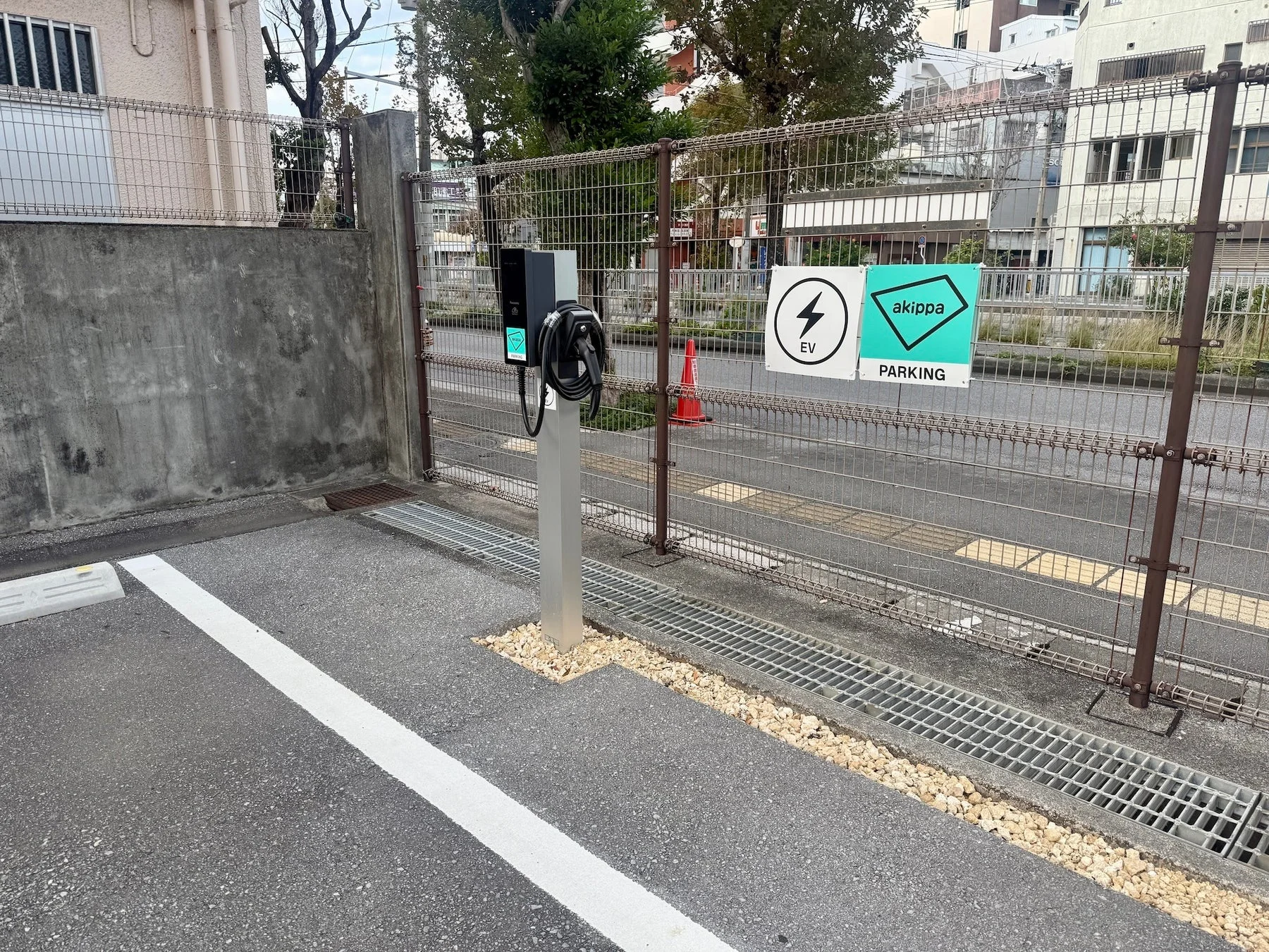akippaのEV充電ステーションが設置された駐車スペースの風景。フェンス越しに道路や建物が見える、都市部または郊外の一角を捉えています。