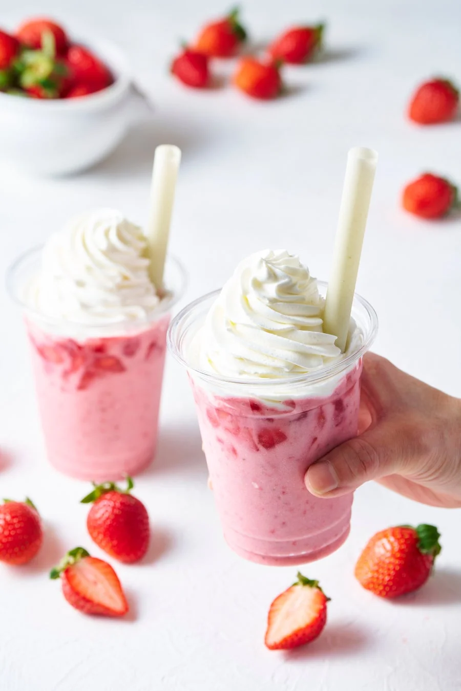 白い背景に、ホイップクリームと太いストローが添えられた2つのいちごミルクドリンクが写っています。周りには新鮮ないちごが散らばっており、手がいちごミルクのカップを一つ持っています。