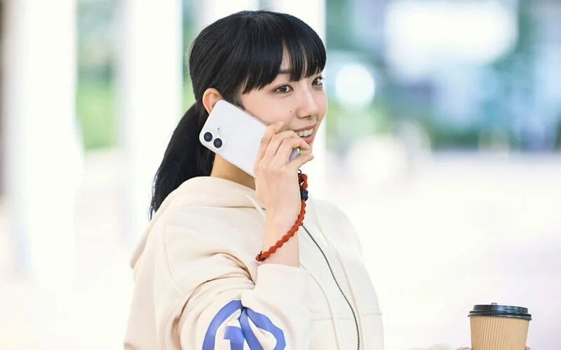 ポニーテールの若い女性が笑顔でスマートフォンで通話しており、もう片方の手にはコーヒーカップを持っています