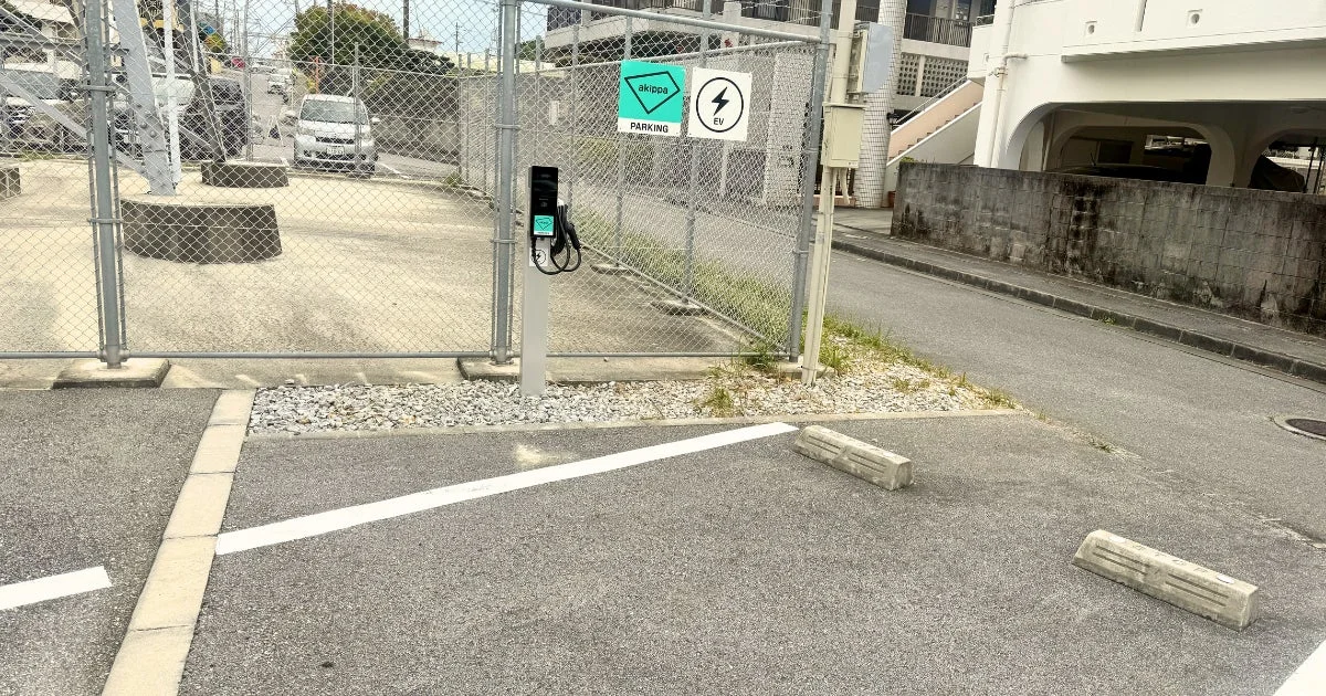 屋外の駐車場に設置されたEV充電ステーションの様子です。akippaの駐車スペースであることを示す看板とEV充電の表示があります。奥にはフェンスがあり、白い車が見えます。