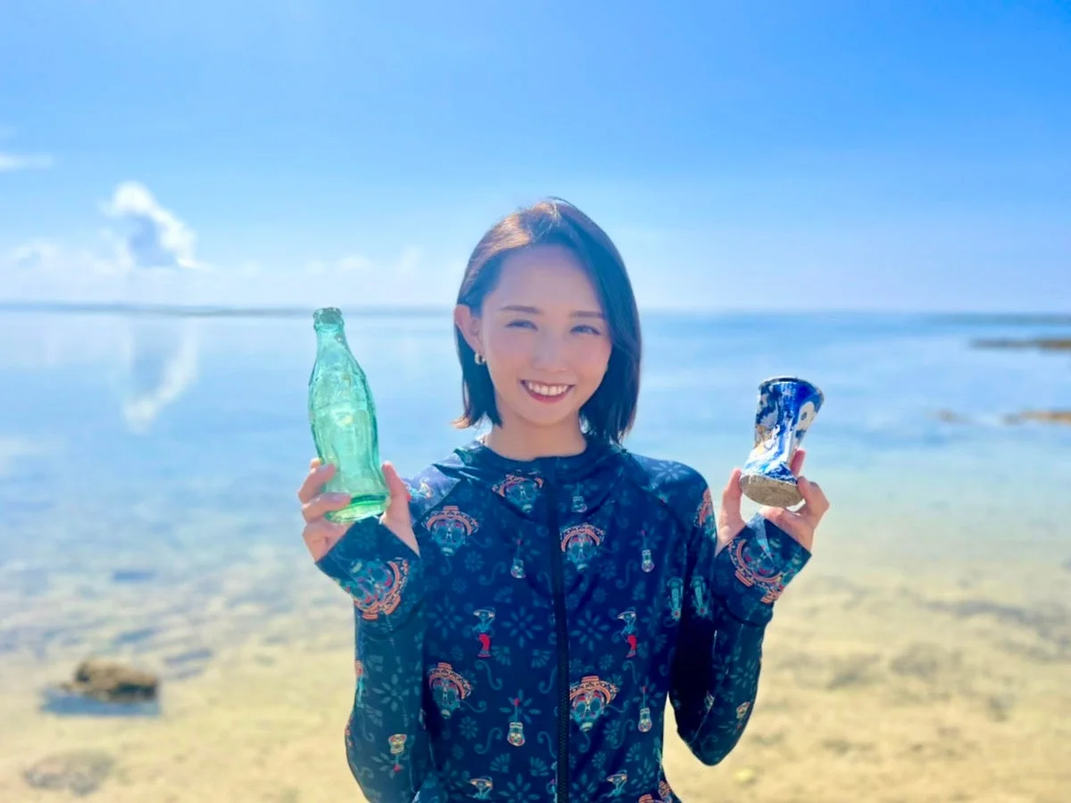 青い空と海を背景に、ビーチで緑色のボトルと青い陶器の破片を手に持ち笑顔で立つ東真七水氏