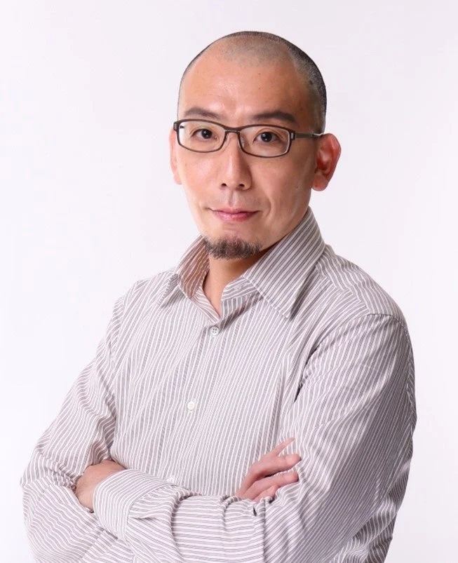 白い背景の前で、メガネをかけ、坊主頭とヤギひげの男性がストライプシャツを着て腕を組んでいます。真剣な表情でカメラを見つめています。