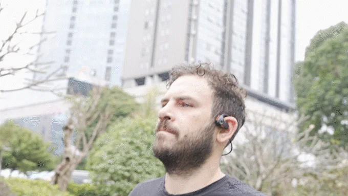 ひげを生やした男性がイヤホンを装着し、屋外で何かを見上げている様子です。