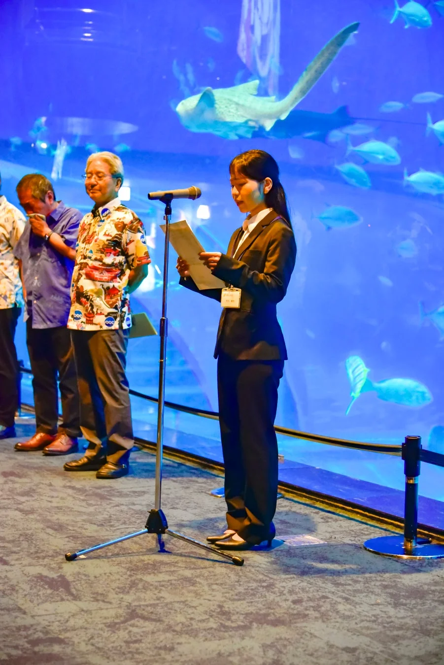 水族館の大水槽を背景にマイクでスピーチをするスーツ姿の女性