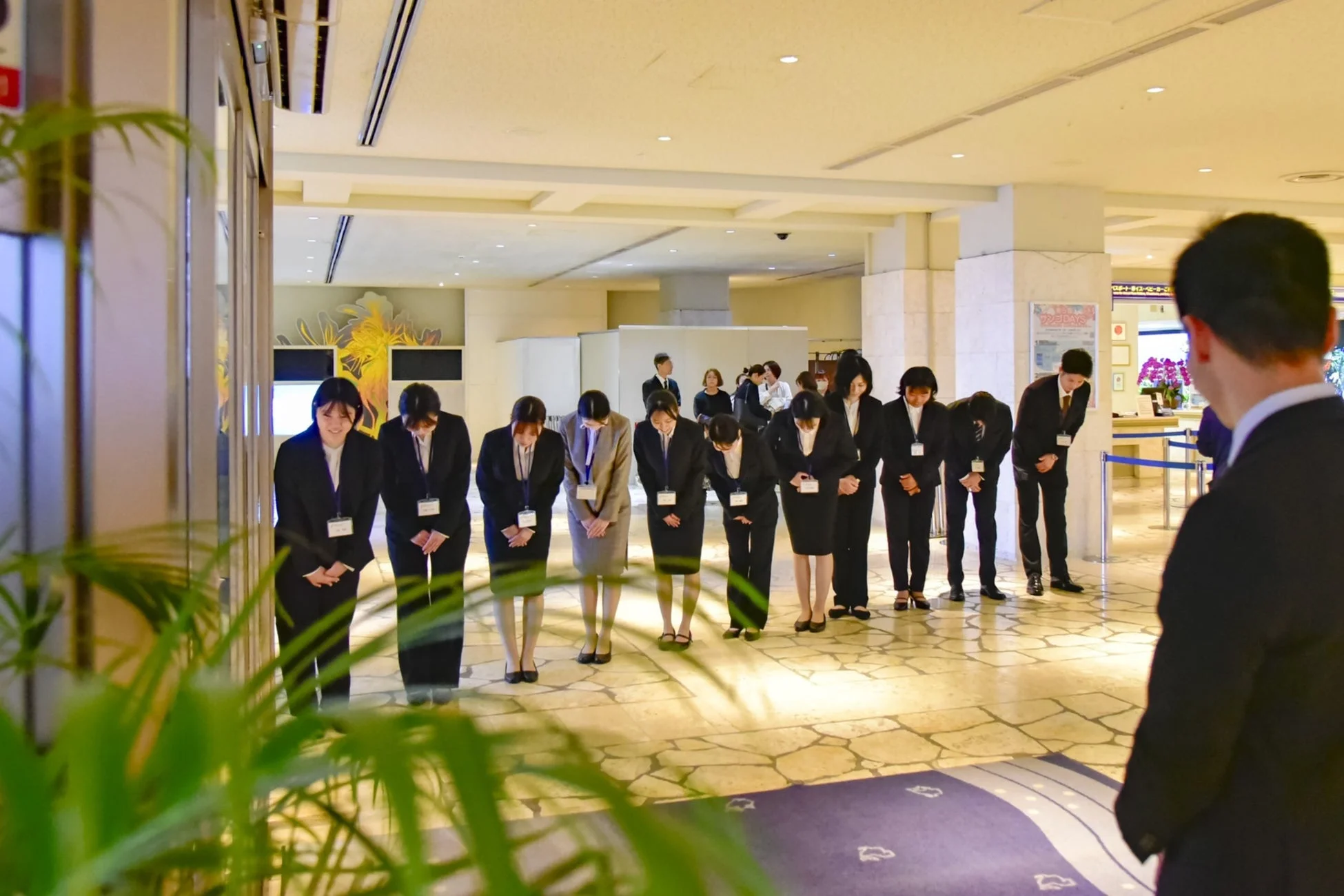 ホテルロビーのような場所で、スーツ姿のスタッフが一斉に深々と頭を下げてお辞儀をしている場面