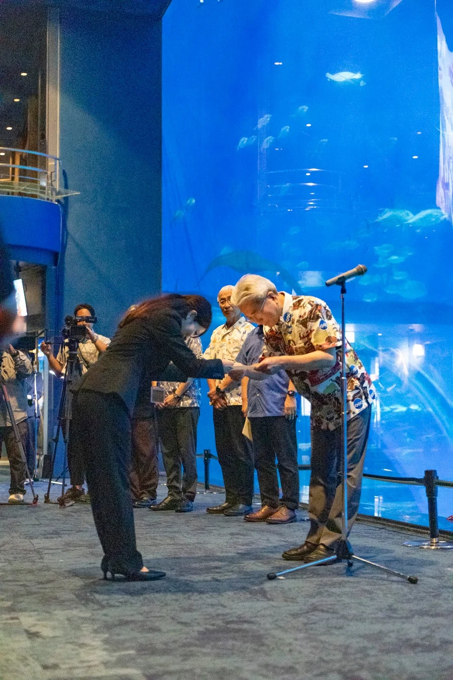 水族館の大水槽の前でアロハシャツを着た男性にお辞儀をするスーツ姿の女性