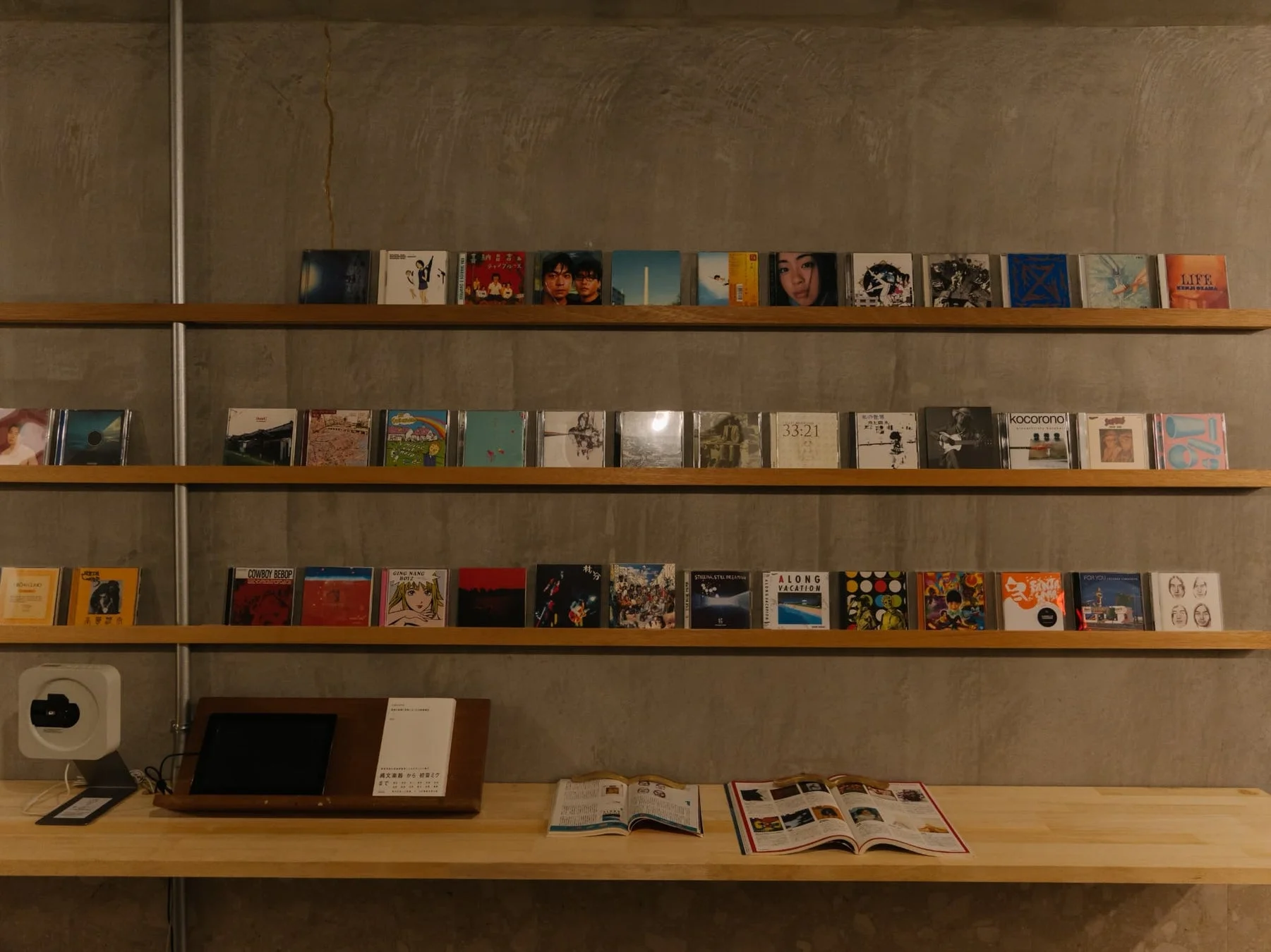コンクリート壁に多数のCDアルバムが並ぶ棚と、タブレットや書籍が置かれたデスク