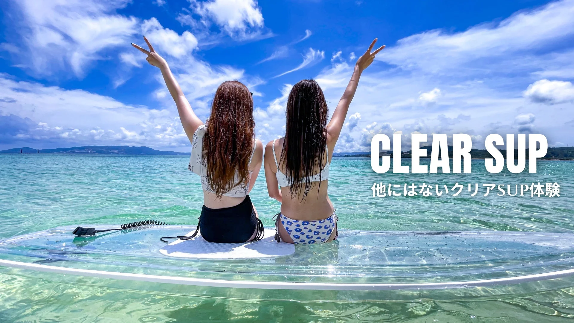 透明なサップボードに乗った二人の女性が、美しい青い海と空の下でピースサインをしており、特別なマリンスポーツ体験を楽しんでいる様子
