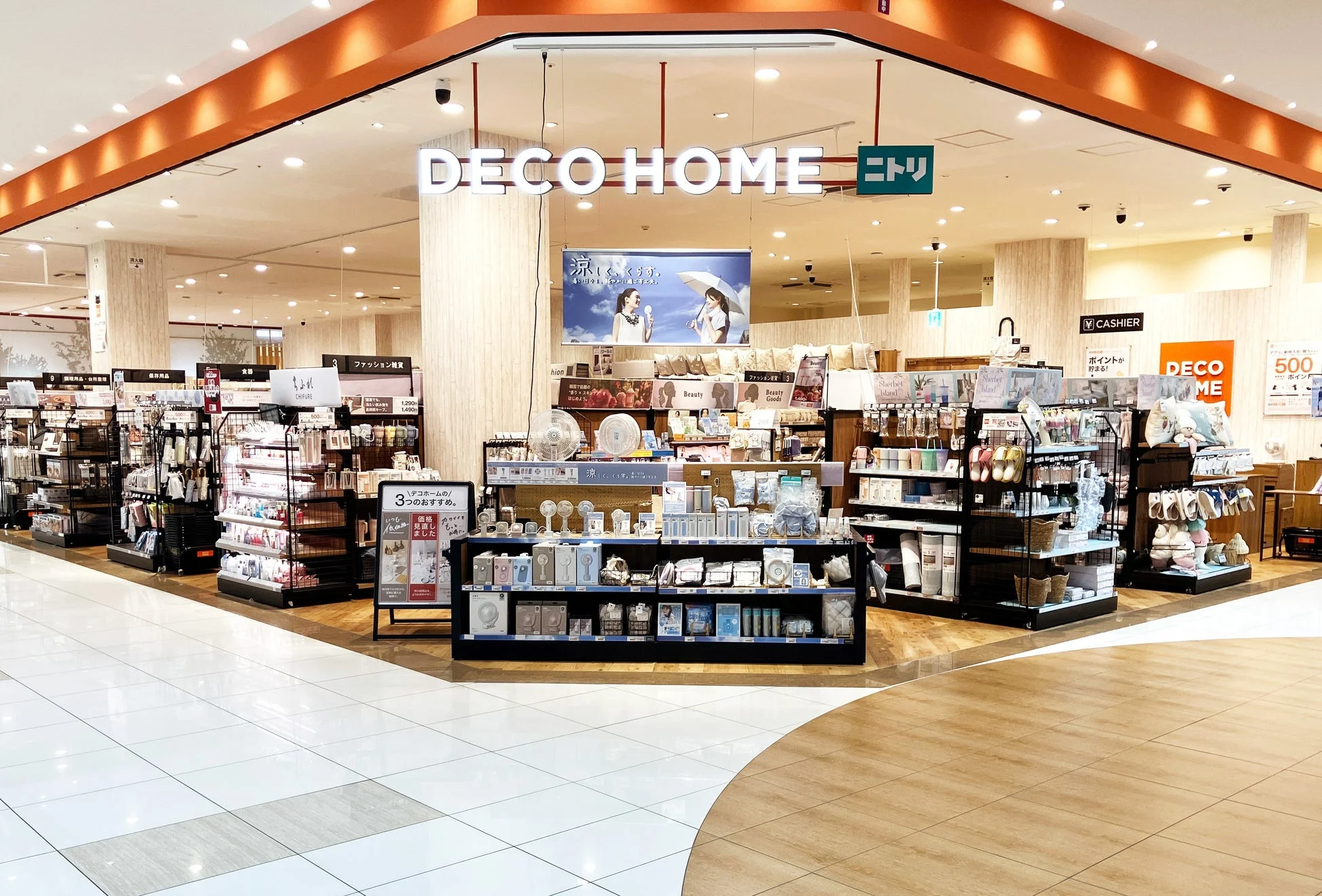 ニトリグループが展開する生活雑貨店「デコホーム」の店内風景です。小型扇風機などの夏物商品や、ファッション雑貨、化粧品、キッチン用品など、幅広い商品が整然と陳列されています。