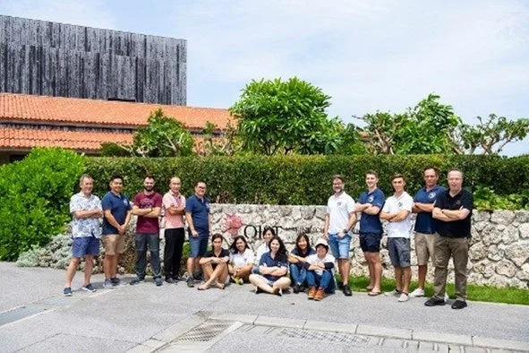 沖縄科学技術大学院大学(OIST)の建物前で、多様なメンバーが集合写真を撮っています。晴れた日の屋外で、リラックスした雰囲気が伝わります。