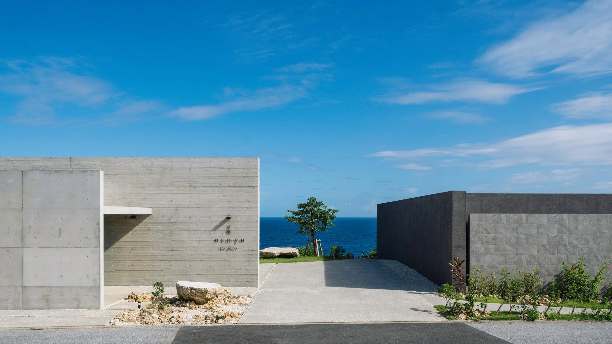 青い海と空が広がる開放的な空間に、モダンなコンクリート造りの建物が佇んでいます。シンプルなデザインの建物の間から海が見え、手前には岩や植物が配置された、静かで美しい風景です。