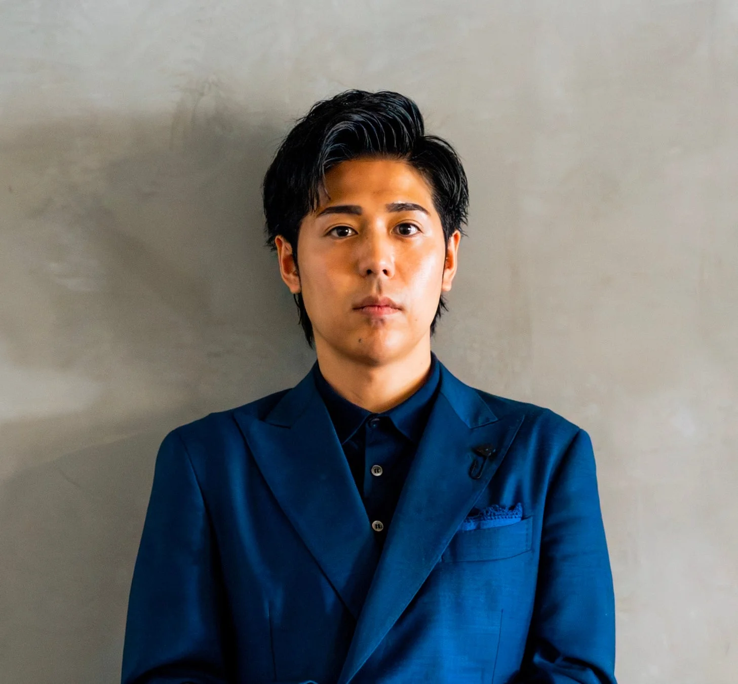 青いスーツを着た若い男性が、グレーの壁を背景にカメラをまっすぐ見つめているポートレート写真です。落ち着いた雰囲気で、ビジネスシーンやプロフィール写真に適しています。
