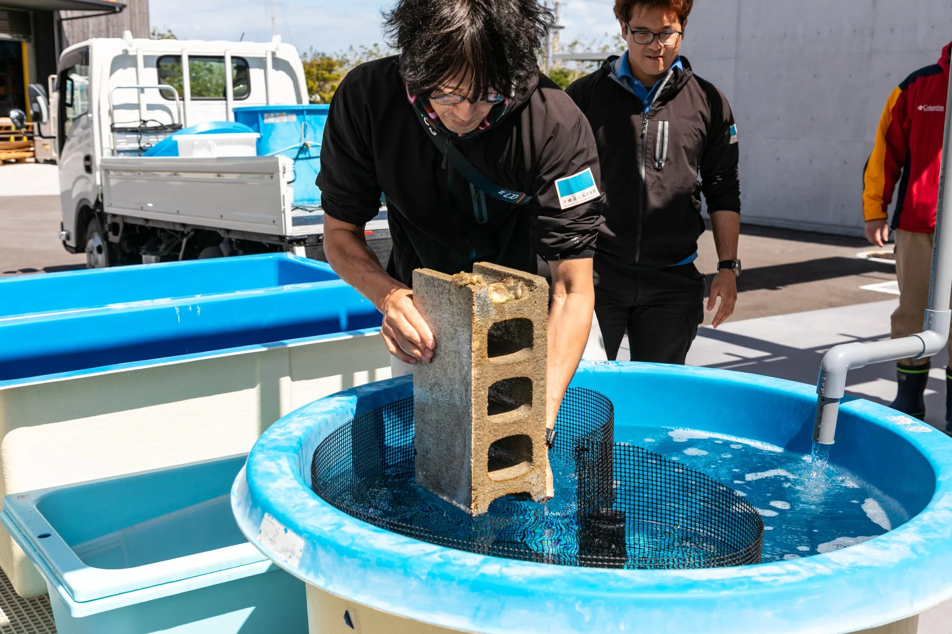 屋外の施設で、男性が水槽にコンクリートブロックを慎重に入れている様子。水生生物の飼育や研究に関連する作業を行っており、別の男性がそれを見守っている。沖縄美ら海水族館のスタッフまたは研究者による作業風景と推測される。