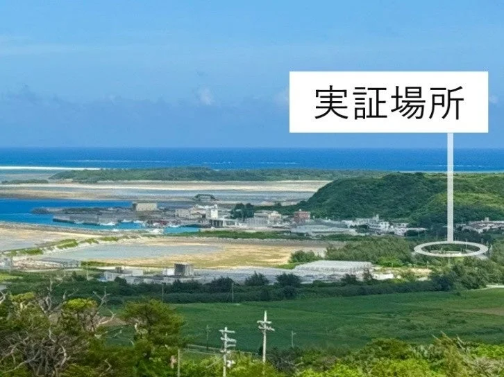 青い空と海が広がる海岸線の風景で、港や小さな島々が見えます。画像中央には「実証場所」と書かれたマーカーがあり、特定の場所を指し示しています。