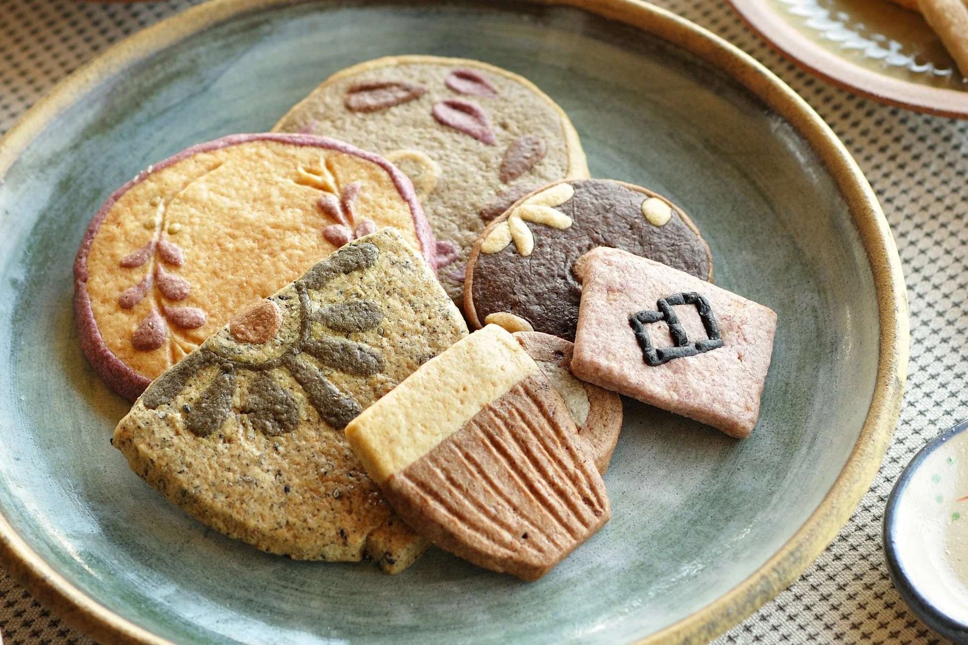 様々な色と形のクッキーが、おしゃれな陶器の皿に美しく盛り付けられています。葉っぱやカップ、独特の模様が施された手作り感あふれる焼き菓子で、お茶請けに最適です。