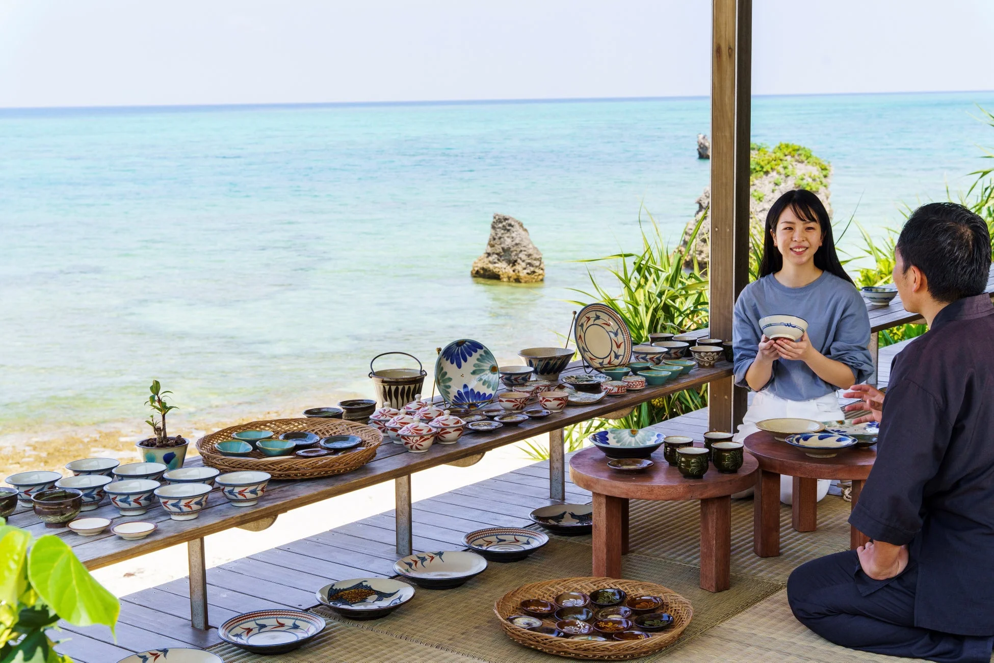 美しいターコイズブルーの海を背景に、屋外のウッドデッキで陶器が多数並べられています。女性が男性に器を見せながら説明しており、沖縄のやちむん店のような雰囲気です。