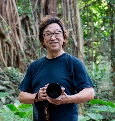 自然豊かな屋外で、黒いTシャツを着たアジア人男性がカメラを手に笑顔で立っています。背景には大きな木々が見え、リラックスした雰囲気が伝わってきます。