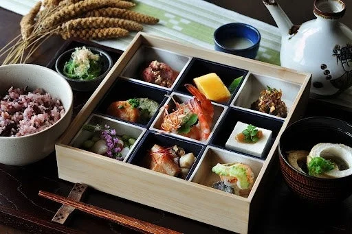 木製の弁当箱に彩り豊かな和食の御膳が並べられています。多種類の小鉢料理、雑穀米、汁物、そして徳利と盃が添えられ、伝統的な日本食の美しさと健康的な食事が表現されています。
