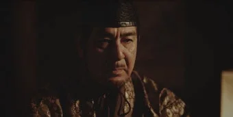 暗い背景の中、伝統的な衣装と黒い帽子を身につけた、髭を生やした厳粛な表情の高齢男性