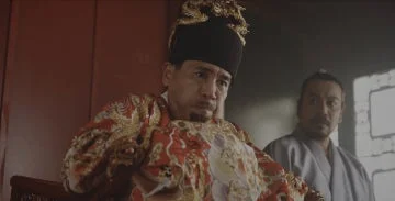 豪華な伝統衣装を身につけた男性が、何かを思案しているような表情で座っています。